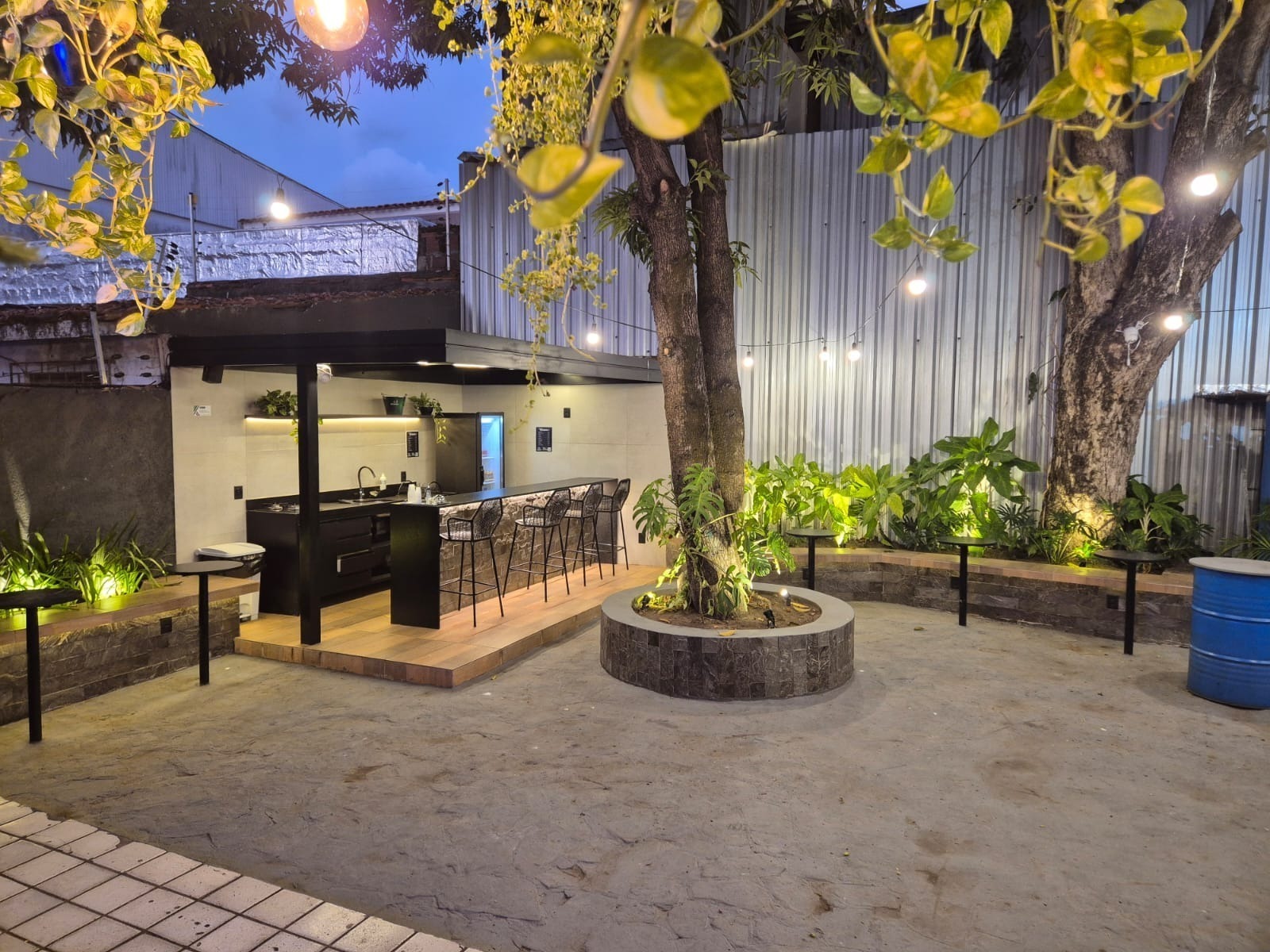 Ambiente gourmet ao entardecer, com bar, bancada, banquetas e a árvore iluminada no meio do pátio