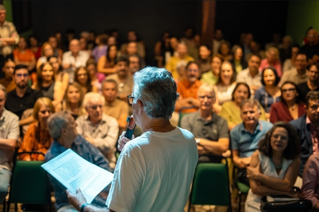 Palestrante de costas, com o microfone na mão, diante de uma plateia lotada no auditório